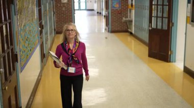 Olgun bir kadın elinde kitap ve kağıtlarla parlak bir okul koridorunda duruyor. Renkli bir atkı takıyor ve yürürken kendinden emin görünüyor..