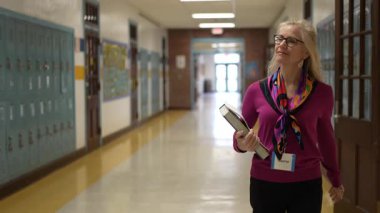 Renkli bir eşarp ve süveter giymiş olgun kadın öğretmen portresi okul koridorunda geziniyor. Kitapları tutuyor ve yoğun bir eğitim ortamında işine dalmış gibi görünüyor..