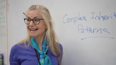 Orta yaşlı bir kadın öğrencilere, beyaz bir tahtanın önünde dururken kalıtsal kompleks kalıpları öğretiyor. Dersini etkili bir şekilde yönlendirmek için notlar kullanıyor..