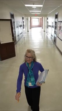 Gözlüklü orta yaşlı bir kadının okul koridorunda dolap ve Amerikan bayrağıyla dolu bir şekilde yürürken çekilmiş dikey videosu kitap ve öğretim materyalleri taşıyor..