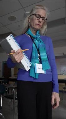 Yetişkin bir kadının boş bir sınıfta elinde bir klasör ve kalem tutarak öğretmenlik sorumluluklarına hazırlandığı dikey bir video. Odaklanmış tavrı eğitime olan bağlılığını yansıtıyor..