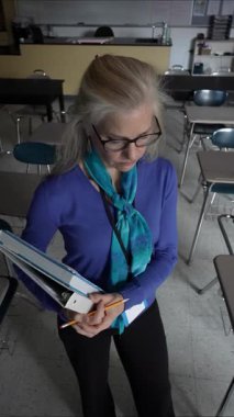 Orta yaşlı, üzgün bir kadının boş bir sınıfta elinde öğretmenlik malzemeleriyle dikey videosu. Gözlük takıyor ve boynunda renkli bir atkı olan mavi bir bluz..