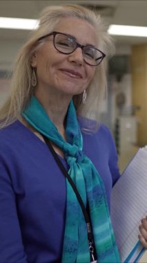 Yetişkin bir kadının boş bir sınıfta elinde bir klasör ve kalem tutarak öğretmenlik sorumluluklarına hazırlandığı dikey bir video. Odaklanmış tavrı eğitime olan bağlılığını yansıtıyor..