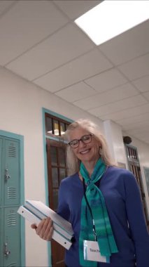 Orta yaşlı kadın öğretmenin elinde birden fazla dosya tutarken gülümsediği dikey bir video. Dolaplar duvarlarla çevrili ve bir Amerikan bayrağı karşılayıcı bir atmosfer yaratıyor..