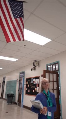 Gözlüklü orta yaşlı bir kadının okul koridorunda dolaplarla süslenmiş bir şekilde yürürken çekilmiş dikey videosu bir defter taşıyor. Arka planda bir Amerikan bayrağı asılı.
