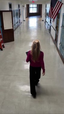 Orta yaşlı öğretmenin kameradan uzaklaştığı dikey bir video ve elinde tablet olan boş bir okul koridoru. Duvarlar dolap ve geniş pencerelerle kaplı..