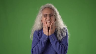 50 'li yaşlarında çenesine dokunarak rahatsızlık belirtileri gösteren olgun bir kadın. Uzun, dalgalı saçları var ve gözlük takıyor. Canlı yeşil bir ekranın karşısında duruyor..