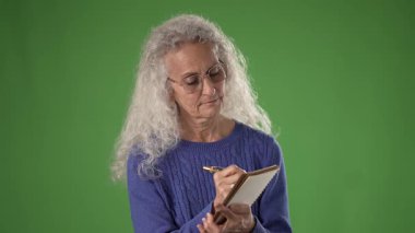 50 'li yaşlarda, uzun gri saçlı olgun bir kadın derin düşüncelere dalmış elinde bir not defteri ve kalemle yeşil perde arkasına yaslanıyor. İfadesi düşüncelere daldığını gösteriyor..