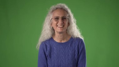 50 'li yaşlarda, kıvırcık gri saçlı orta yaşlı bir kadın yeşil ekran karşısında rahat mavi bir kazak giyerken sevinçle bağırıyor, muhtemelen eğlenceli bir proje için..