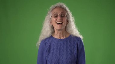 50 'li yaşlarda, kıvırcık beyaz saçlı olgun bir kadın mavi bir kazak giyer ve yeşil ekranın önünde duran seyirciyi büyük bir gülümsemeyle selamlar..