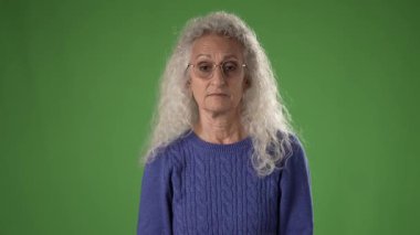 50 'li yaşlarında, kıvırcık saçlı, gözlüklü, hareketli, kafası karışmış bir kadın. Parlak yeşil bir ekranın önünde duygularını vurguluyor..