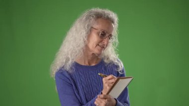 50 'li yaşlarında, elinde not defteri ve kalemle gülümseyen kıvırcık gri saçlı olgun bir kadın. Yeşil ekranda yazdığı gibi meşgul ve mutlu görünüyor..