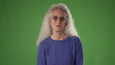 50 'li yaşlarda, gri saçlı ve gözlüklü bir kadın yeşil bir ekranın karşısında dururken yürekten duygular taşır. Samimi bir yansımayla meşgul gibi görünüyor..