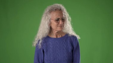 50 'li yaşlarında, uzun gri saçlı bir kadın şaşkınlıkla parmağını kaldırıyor. Mavi bir kazak giyiyor ve gözlüğü var. Arka plan parlak bir yeşil..