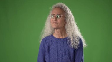 Uzun kıvırcık saçlı olgun bir kadın mavi bir kazak giyerken şşş hareketi yapar. Parlak yeşil arka plan onun ifadesini geliştirir, ana odaklanır..
