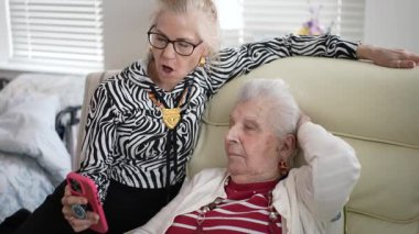 Yaşlı bir kadın koltukta dinlenirken kızı ona telefonda bir şey gösteriyor, rahat bir ortamda görüntülü bir sohbet üzerinden bağlanıyor..