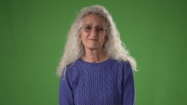 50 'li yaşlarda olgun bir kadın tarafsız bir ifadeyle ayakta duruyor. Dalgalı gri saçları var ve parlak yeşil bir ekranın önünde dururken gözlük takıyor..