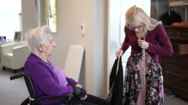 Bir kadın, tekerlekli sandalyede oturan yaşlı akrabasıyla sohbet eder. Kadın kıyafetleri sınıflandırıyor ve odada yararlı bir atmosfer yaratıyor..