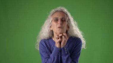 50 'li yaşlarda, mavi bir kazak giyen olgun bir kadın ellerini çapraz parmaklarla tutar, yeşil bir ekranda heyecan ya da beklenti gösterir..