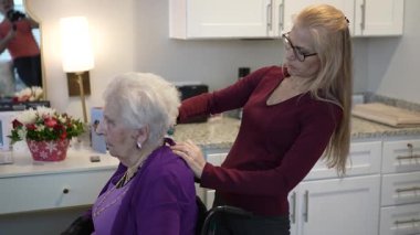 Bakıcı, yaşlı bir kadının saçlarını nazikçe kişisel dokunuşlar ve dekorasyonlarla dolu sıcak ve davetkar bir odada tarar..