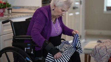 Tekerlekli sandalyedeki yaşlı kadın, parlak mutfağında oturmuş, pratik becerilerini ve yaratıcılığını sergileyerek çizgili bir kumaşı katlamaya odaklanıyor..