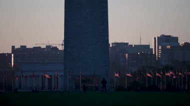 Washington Anıtı, rüzgarda dalgalanan sayısız Amerikan bayraklarıyla altın bir günbatımına karşı dimdik ayakta duruyor. Arka planda Lincoln Anıtı görülebiliyor ve şehir manzarası daha da uzanıyor..