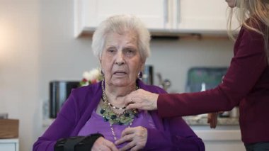 Mor bir hırka giyen yaşlı bir kadın, birisi ona güzel bir kolye takmasına yardım ederken içeride oturuyor. Atmosfer sıcak ve destekleyici..