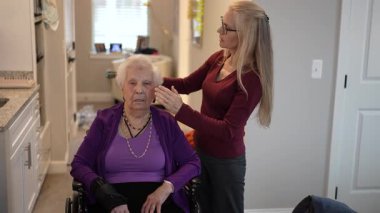 Sıcak bir mutfakta, olgun bir bakıcı tekerlekli sandalyede oturan yaşlı bir kadının saçını şekillendirir. Etkileşimlerinde şefkat ve özen gösterir..