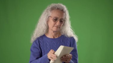 50 'li yaşlarda, uzun, gri saçlı orta yaşlı bir kadın mektup okurken gülümsüyor ve şaşırıyor. Mavi bir kazak giyiyor ve yeşil bir ekranın önünde duruyor..