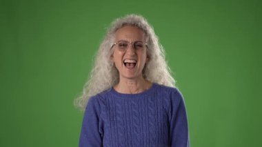 50 'li yaşlarda kıvırcık gri saçlı olgun bir kadın mavi bir kazak giyerken neşeyle gülümsüyor. Arka plan canlı yeşil, bir stüdyo ortamını gösteriyor..