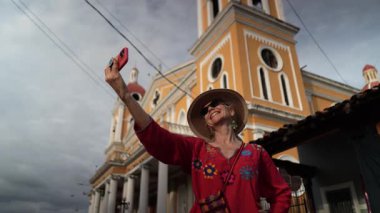 Granada, Nikaragua - 01 13 2025: Olgun bir kadın Granada, Nikaragua 'da canlı bir kilisenin önünde neşeli bir ifadeyle selfie çekiyor. Mimari ve giyimi yerel kültürü yansıtıyor..