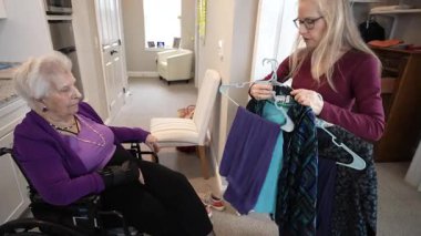 Tekerlekli sandalyedeki yaşlı bir kadın, kızı tarafından sunulan kıyafet seçeneklerini inceleyerek, sıcak bir ev ortamında aile bağlarını ön plana çıkarıyor..