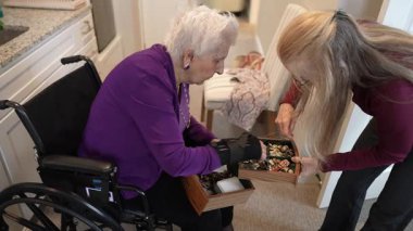 Tekerlekli sandalyedeki olgun bir kadın ve kızı sıcak ve davetkar bir şekilde mücevher kutularını karıştırıyorlar. Gülümsemeleri ve bağlantı anlarını paylaşıyorlar..