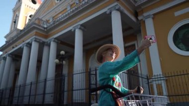 Olgun bir kadın, Granada, Nikaragua 'da canlı bir katedralin yakınında bisiklet sürerken selfie çeker ve güneşin altındaki neşeli turistik deneyimini yakalar..