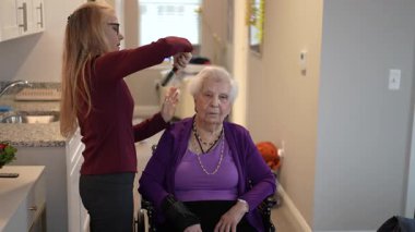 Tekerlekli sandalyedeki yaşlı kadın sakince oturur. Ailesi hafifçe saçlarını tarar. Işıl ışıl bir mutfak alanında..