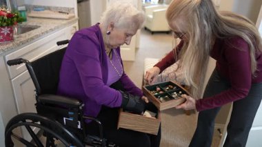 Tekerlekli sandalyedeki yaşlı bir kadın mücevherleri parlak, çağdaş bir yaşam alanında bir bakıcının yardımıyla ayırıyor..