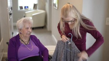 Bir bakıcı, yaşlı bir kadına tekerlekli sandalyede yardım ederken onlar da kişisel dokunuşlarla dolu, aydınlık, hoş bir yaşam alanında etkinlik hazırlıyorlar..