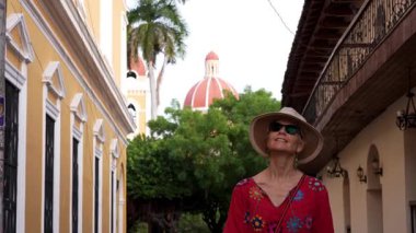 Granada, Nikaragua - 01 13 2025: Güneş gözlüklü ve hasır şapkalı olgun bir kadın renkli mimari ve yemyeşil ile çevrili Granada, Nikaragua 'nın canlı sokaklarında yürüyor.