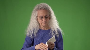 50 'li yaşlarda orta yaşlı bir kadın, düşüncelere dalmış yeşil perde önünde odaklanmış ifadesini sergilerken bir belgeyi inceliyor..