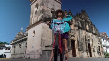 Olgun bir kadın bisikletiyle Granada, Nikaragua 'daki La Merced Kilisesi' nin önünde poz verirken gülümsüyor. Parlak mavi gökyüzü ziyaretinin neşe dolu atmosferine ekleniyor.