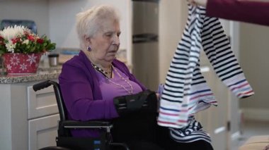 Yaşlı bir kadın tekerlekli sandalyede oturuyor, çamaşırları dikkatlice katlıyor. Parlak ve davetkar bir mutfak alanı, ev dekoru ve yumuşak ışıklandırmayla çevrili..