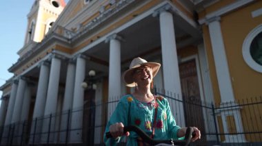 Olgun bir kadın, Orta Amerika 'nın canlı atmosferini yakalamak için Granada, Nikaragua' daki katedralin dışında bisiklet sürerken neşeyle güler..