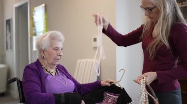 Olgun bir kadın tekerlekli sandalyede oturur, çeşitli inci kolyelerini incelerken ilgili bir görevli de rahat bir yerde en iyilerini seçmesine yardım eder..