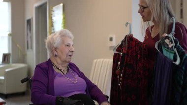 Tekerlekli sandalyedeki olgun kadın başka bir kadının parlak ve modern bir odada tuttuğu renkli elbiseleri gözlemler. Birlikte seçenekleri incelerken atmosfer sakin..