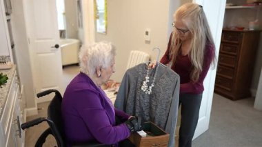 Tekerlekli sandalyedeki yaşlı bir kadın, rahat bir evde giysi ve kişisel eşyaları düzenlerken kızından yardım alır..