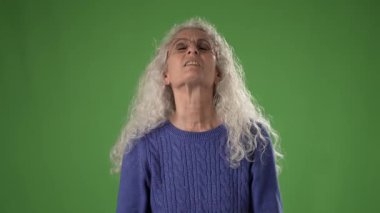 50 'li yaşlarda kıvırcık beyaz saçlı, mavi bir kazak giyerken yukarı doğru hareketler yapan olgun bir kadın. Yeşil ekranın önünde şaşkınlık ifadesi sergiliyor..