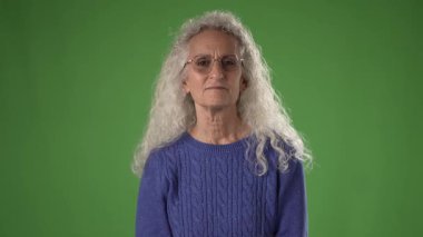 50 'li yaşlarda orta yaşlı bir kadın, elini kulağına kaldırırken gülümsüyor, bu da onun dikkatlice dinlediğini gösteriyor. Kıvırcık gri saçları ve gözlükleri mavi süveterini tamamlıyor..