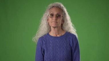 50 'li yaşlarda, kıvırcık gümüş saçlı ve gözlüklü orta yaşlı bir kadın, sakin bir şekilde yeşil ekran karşısında duruyor, düşünceli ifadesini ve mavi kazağını gösteriyor..