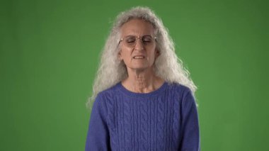 50 'li yaşlarda, mavi bir kazak giyen olgun bir kadın, elini ağzının yanına koyup düşünceli bir jest yaparken yeşil bir arka plan karşısında duruyor..