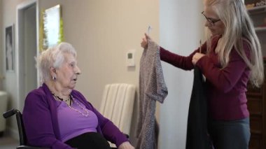 İyi ışıklandırılmış bir odada, tekerlekli sandalyedeki yaşlı bir kadın başka bir kadının tuttuğu iki kazağı inceliyor. Atmosfer rahat ve sıcak, kişisel bir seçim anını vurguluyor..